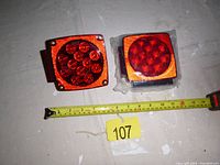 Two square LED trailer tail lights, one loose and one in plastic packaging, side-by-side with measuring tape showing size.