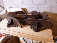 Five dark solid wood shelves stacked with intricately carved decorative brackets on a light wood surface in front of a concrete block wall.