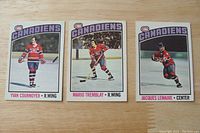 Front view of three Montreal Canadiens hockey cards of Yvan Cournoyer, Mario Tremblay, and Jacques Lemaire displayed on wooden surface.