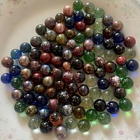 Overhead photo showing a wide assortment of vintage glass marbles with speckled patterns in dark colors, arranged loosely on a floral plate.