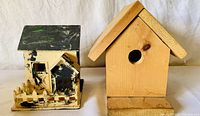 Two wooden bird houses side by side on a white background. The left one painted black and white with detailed front resembling a small house with fence. The right one natural wood with sloped roof and circular entrance hole.