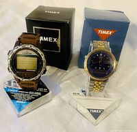 Two Timex men's watches on display stands with original boxes behind each. Left watch is Timex Expedition digital compass with brown leather strap, right watch is gold and silver tone stainless steel analog-digital watch with blue dial.