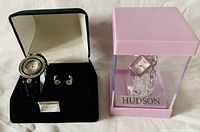 Photo showing the Elizabeth Taylor Euro Bracelet watch with black strap and matching earrings in a black velvet box, positioned beside the Hudson watch in its pink clear display box.