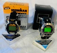 Two Timex Ironman Triathlon watches on stands with their original boxes behind. The black watch has black resin strap, orange label, and data-link text on strap, black, orange and yellow face. The steel watch has round stainless steel body, digital green display, black strap with the original black Timex box behind.