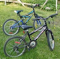 Two youth bicycles outdoors on grass, one black and one blue, showing side and rear views of both bikes, tires and seats visible.