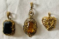 Three antique filigree pendants with colored stones laid out on a white fabric background: rectangular dark stone pendant, amber stone filigree pendant, and heart-shaped filigree pendant with a flower.