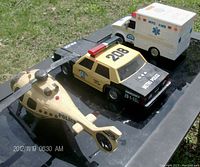 Photo showing Metro Police car, Police helicopter, and NYC EMS ambulance toy models on a black surface outdoors with clear details and markings.