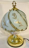 Full view of the table lamp showing white glass shade with gold floral designs and brass-colored metal base.
