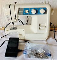 Side angle photo of the Brother VX-1140 sewing machine with foot pedal and accessories including multiple metal bobbins in plastic bags in the foreground.