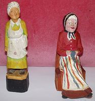 Front view of two wooden figurines against a pink background, showing a standing elderly woman with apron in yellow dress and a seated elderly woman in red top and striped skirt on a wooden chair.