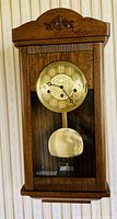 Front view of solid oak Westminster wall clock with visible carved decoration and brass pendulum inside a glass door.
