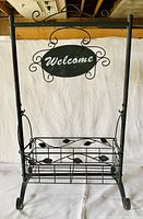 Full view of black metal plant stand with hanging "Welcome" sign, scrollwork, and leaf detailing on basket holder and sides.