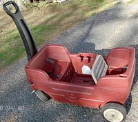 Side angle view of the red plastic Step2 wagon with a gray handle, showing the seating area with seat belts and wheels, on an outdoor pavement.