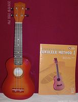 Front view of soprano ukulele with sunburst finish and instructional manual standing next to it.