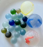 Photo showing 20 sea glass marbles on a white surface, colors include blue, green, yellow, white and olive with frosted texture.