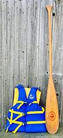 Photo showing wooden canoe paddle and blue-yellow life jacket against wooden plank background