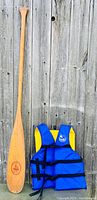 Wooden paddle and blue/yellow PFD placed vertically against a wooden fence, showing full length of paddle and folded PFD.