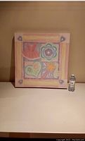 Front view of the square colorful kids wall art with dress, flower, heart, and star relief designs, sized 24x24 inches, placed on a table beside a water bottle for scale.