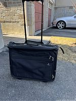 Front view of black Atlantic garment bag with telescoping handle extended, showing exterior zippered compartments and handle.