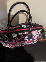 Front view of colorful floral patterned Barbie handbag with black handles and Barbie logo on the front pocket area.
