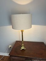Full view of antique brass church lamp standing on a wooden surface, showing tall slender body, footed base with ornate raised face details, and beige fabric shade illuminated.