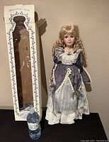 Porcelain doll next to its decorative box and water bottle for size reference, showing doll's front and dress detail.