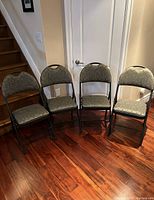 Four folding chairs arranged in a row against a wall, showing cushioned seats and backs in gray patterned fabric with black metal frames.
