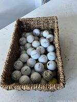 Top-down image of a rectangular woven basket filled with used golf balls, mostly white with one yellow ball visible. Basket placed on a white surface.