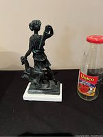 Side view of the Greek statue depicting a standing hunter with dog on white marble base, placed next to a bottle for size reference.