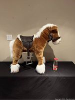 Side view of the mechanical horse plush toy showing the brown body, white mane and tail, and attached black saddle and harness. The toy is placed on a black surface with a bottle nearby for size reference.