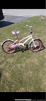 White and pink kids bike standing on grass with training wheels attached and visible chain guard with colorful design.