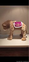 Side profile of the plush lion toy showing tan fur, fluffy mane, and purple embroidered saddle blanket.