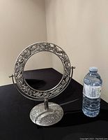 Photo of round vanity mirror with ornate pewter floral frame and magnifying feature, shown on black surface alongside water bottle for scale