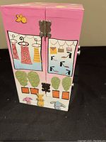Pink rectangular musical jewelry storage box featuring illustrations of clothes, shoes, plants, and cartoon women with pets outside a London boutique