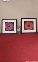 Front view of two square framed pictures of flowers, one pink and one red, placed side by side with a water bottle between them for size reference.