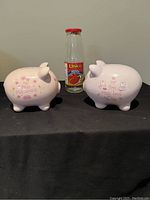 Front view of two pink ceramic piggy banks with printed sayings and a size reference bottle