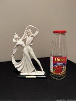 Matte white ceramic figurine of a dancing couple shown next to a standard size glass jar for scale. Figurine appears about 10 inches tall, mounted on a black base with plaque.