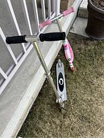 Silver kids scooter with black foam grips and black deck with white design leaning against railing on grass.
