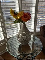 Photo of a woven wooden or bamboo vase with artificial yellow, orange, red, and pink flowers on a round glass table near window blinds.