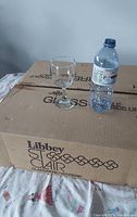 Closed cardboard box labeled Libbey Stemware GLASSWARE/VERRE placed next to a filled water bottle and a sample glass wine glass for size comparison.