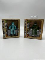 Front view of two StikBot figures side by side in packaging, one teal and one green, showing intact sealed boxes.