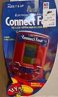 Front of sealed Connect Four electronic handheld game package showing red device with three blue buttons and screen. Packaging is blue with yellow and white text, includes battery and hints at 2 games and 3 skill levels.