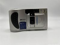 Front angle of silver HP PhotoSmart 315 digital camera showing lens cover, flash, and model details sticker.