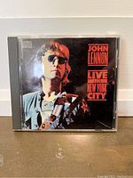 Front of John Lennon Live In New York City CD jewel case, shows John Lennon with guitar and sunglasses, red title text on black background.