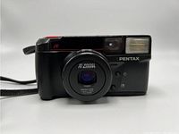 Front view showing lens, built-in flash, autofocus label, Pentax branding and shutter button with wrist strap attached.
