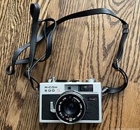 Front view of Ricoh 500 G camera showing lens, body with black grip, model name and attached black neck strap.