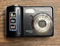 Front view of the Samsung Digimax S600 camera showing lens, flash, and branding with megapixel and zoom info stickers.