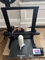 Full view of the Voxelab 3D printer assembled and positioned on a cardboard surface, showing the black aluminum frame, build plate with partially printed white object, and touchscreen control panel.
