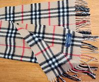 Photos show beige Burberry scarf featuring the iconic black, white, and red check pattern with fringed ends. Tag visible with brand and material details.