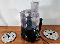 Front angle photo showing the food processor base, clear plastic bowl with black handle, and crinkle cut and fine shred disk attachments next to it.